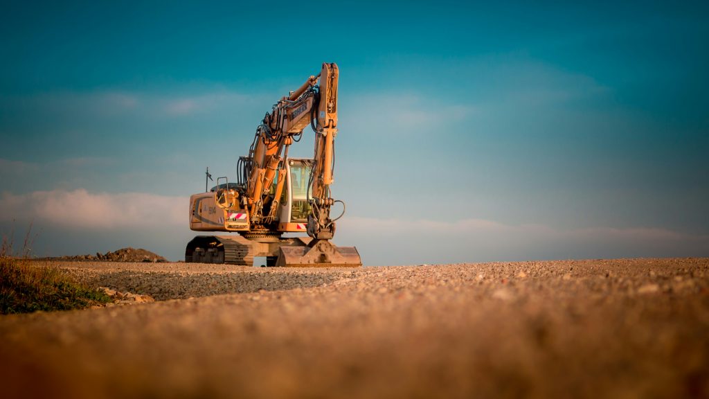 A yellow excavator and an orange soil compactor parked on a construction site with rocky terrain in the background.